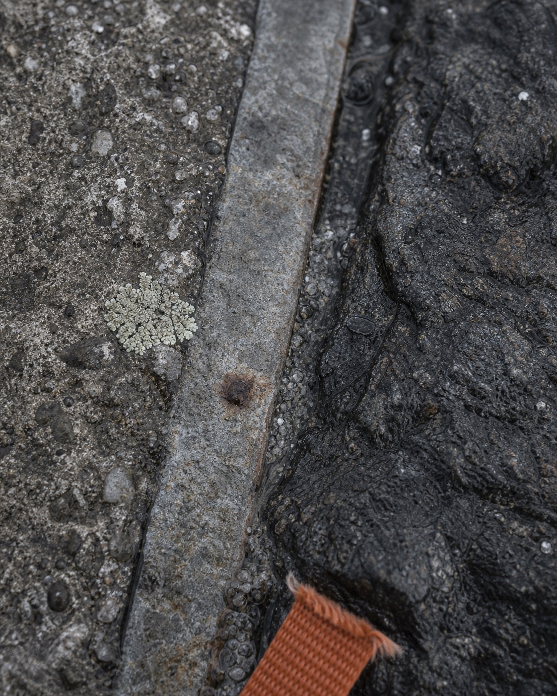 Wet concrete, dark coastal stone, galvanized steel, lichen, and a frayed orange strap in close detail