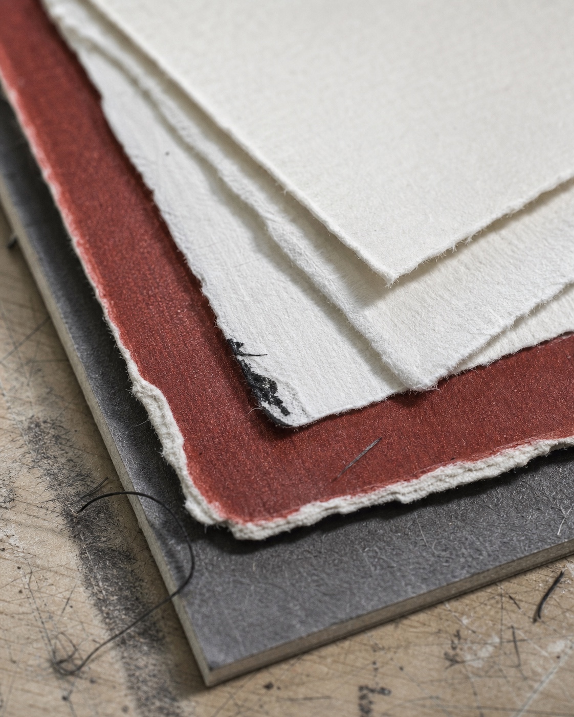 Deckled cream papers over oxide-red stock and graphite board with loose black fibers